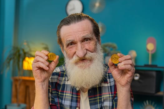 Elderly man freelancer holding two gold BTC coins successful developer programmer. Grandfather stock trader earning bitcoins after online monitoring trading operations. Increasing wealth prosperity