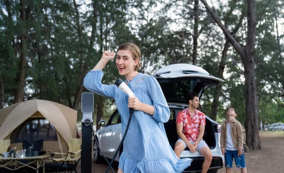 Outdoor adventure and family vacation camping at sea travel by eco friendly car. Cheerful woman or mother holding, pointing EV charger point with playful and happiness posture in campsite. Perpetual