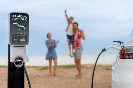 Alternative family vacation trip traveling by the beach with electric car recharging battery from EV charging station with blurred cheerful and happy family enjoying the seascape background. Perpetual