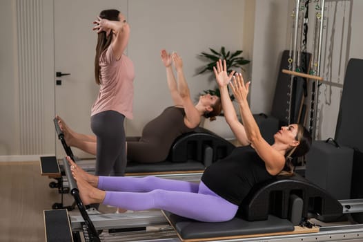 Two pregnant women are doing Pilates on a reformer. The instructor teaches prenatal yoga