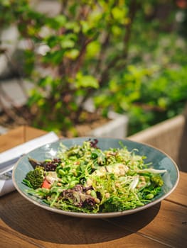 Fresh green salad in plate made of green leaves mix, broccoli and tomatoes on wooden table outdoor restaurant. Variety of healthy fresh harvested eatable greens. Raw food, organic, detox diet concept