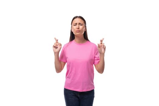 young slim brunette woman in a pink basic t-shirt crossed her fingers on a white background with copy space.