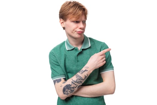 young red-haired man dressed in a green t-shirt points his finger to the side against the background with copy space.