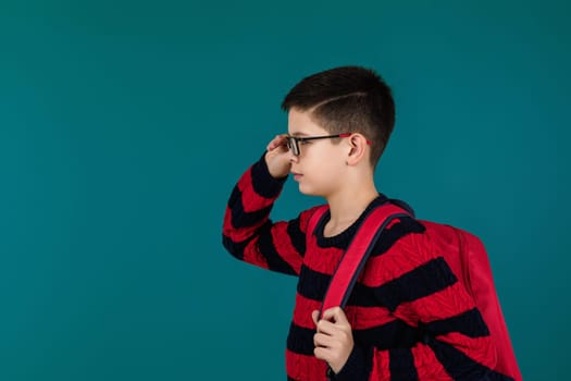 little cheerful school boy wearing glasses with red backpack over blue background. copy space