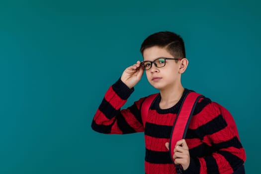 little cheerful school boy wearing glasses with red backpack over blue background. copy space
