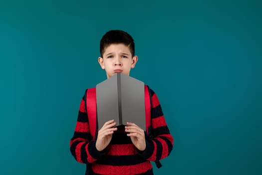 little cheerful school boy reading a book over blue background. child covers his face with book. blank space on book for your text