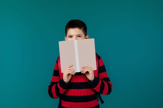 little cheerful school boy reading a book over blue background. child covers his face with book. blank space on book for your text