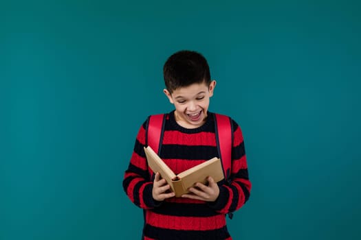 smiling little cheerful school boy reading interesting book over blue background, copy space. School concept.