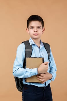 little cheerful school boy with backpack holding a book over beige background