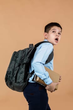 little frightened shocked school boy with backpack holding a book and looking up over beige background