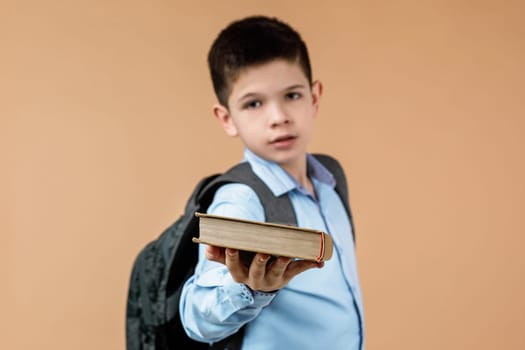 little cheerful school boy with backpack giving book over beige background. focus on book