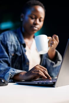 Focused African American freelancer working on her digital laptop while drinking a cup of coffee. Close-up of black woman holding a mug and surfing the net on her personal computer.