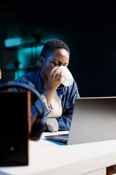 Portrait of african american remote worker drinking coffee while working on her digital laptop. Young black woman with wireless computer on desk is sipping beverage from a cup.