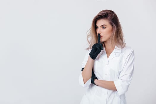 Portrait of young female professional doctor in black gloves shows sign silence. cosmetologist in studio
