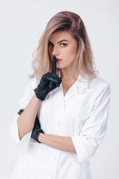 Portrait of young female professional doctor in black gloves shows sign silence. cosmetologist in studio