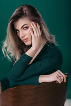 Sensual beautiful blonde woman in green dress sitting on chair and looking at camera against green background