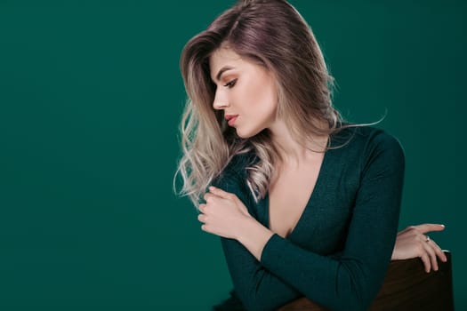 Sensual beautiful blonde woman in green dress sitting on chair against green background