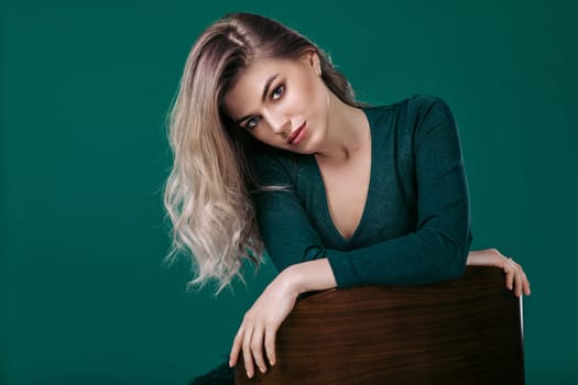 fashion portrait of sensual beautiful blonde woman in green dress sitting on chair and looking at camera against green background