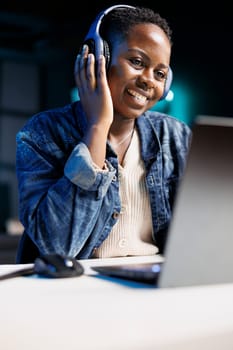 Black woman joyfully using a laptop and wireless headphones. African american female influencer multitasks, attending an online meeting while listening to music in her headset.