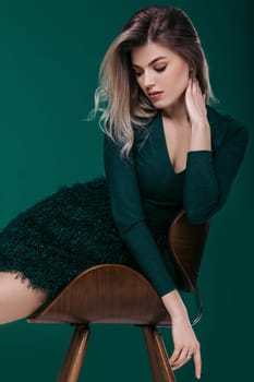 Sensual beautiful blonde woman in green dress sitting on chair against green background