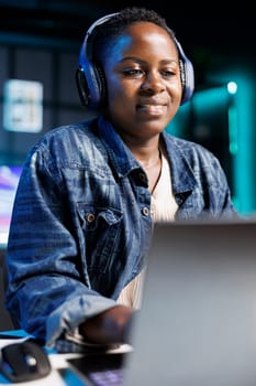 African American freelancer working at home, wearing wireless headphones while browsing the internet and attending a video call. Close-up of smiling black woman looking at her laptop screen.