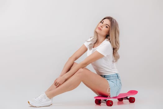 beautiful woman blonde in white t-shirt and denim shorts sitting on pink skateboard