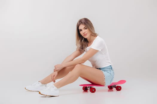 beautiful woman blonde in white t-shirt and denim shorts sitting on pink skateboard