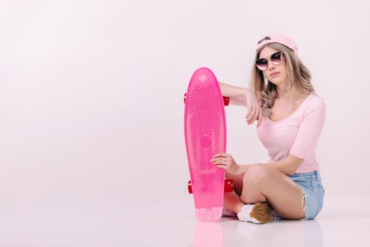 Cute woman in sunglasses, pink t-shirt and cap sitting on the floor with pink skateboard. teenager