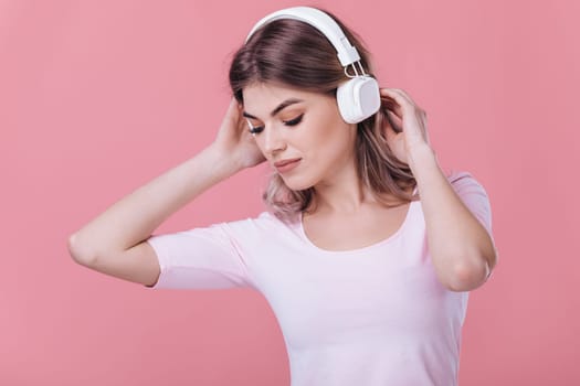 beautiful blonde woman in pink t-shirt and white headphones listens to music on pink background