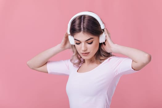 beautiful blonde woman in pink t-shirt and white headphones listens to music and smiling with closed eyes on pink background