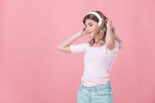 happy young blonde woman in pink t-shirt and white headphones listens to music on pink background. summer studio photoshoot