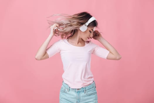 beautiful blonde woman in pink t-shirt and white headphones listens to music and dancing on pink background