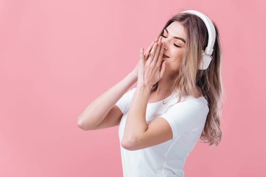 happy young blonde woman in t-shirt and white headphones listens to music on pink background
