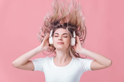 beautiful blonde woman in t-shirt and white headphones enjoying listens to music on pink background. hair fly