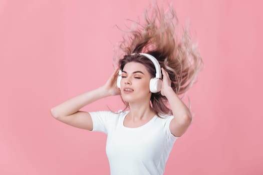 beautiful blonde woman in t-shirt and white headphones listens to music on pink background. hair fly