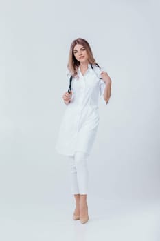Full length portrait of medical physician doctor woman in white coat with stethoscope looks at camera on light background.