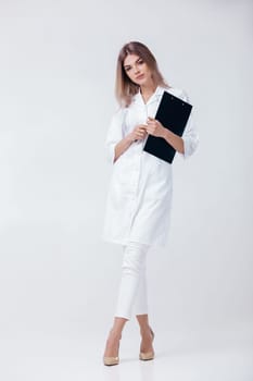 Full length portrait of medical physician doctor woman in white coat holds folder and looks at camera on light background.