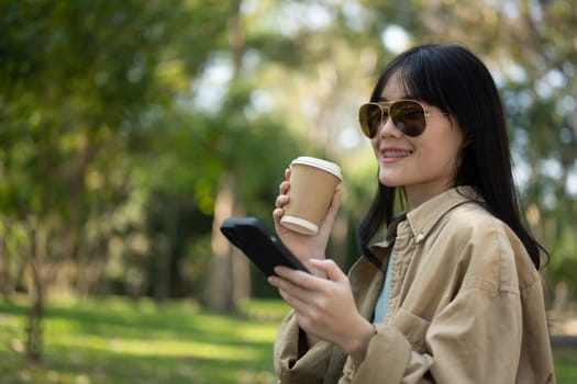 Smiling young woman wearing sunglasses enjoying her takeaway coffee and using mobile phone in the park.