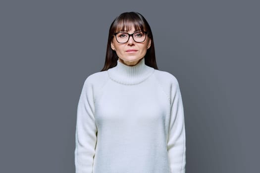 Middle-aged beautiful serious woman in white sweater on gray studio background. Confident mature female in glasses looking at camera. Beauty, lifestyle, business, work, 40s people concept