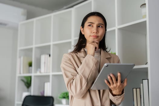 young asian woman, company worker in suit, smiling and holding digital tablet, standing over working space background.