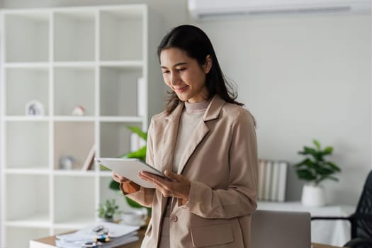 young asian woman, company worker in suit, smiling and holding digital tablet, standing over working space background.