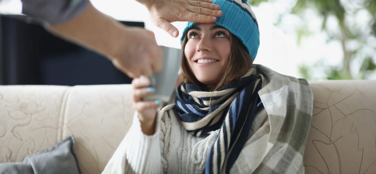 Man hand touching forehead of sick woman in hat and giving cup of tea. Treatment of seasonal colds at home concept