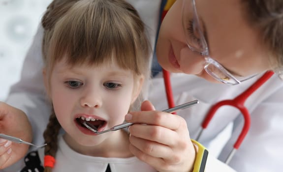 Dentist examining teeth of little girl with steel instruments in clinic. Annual dental checkups for children concept
