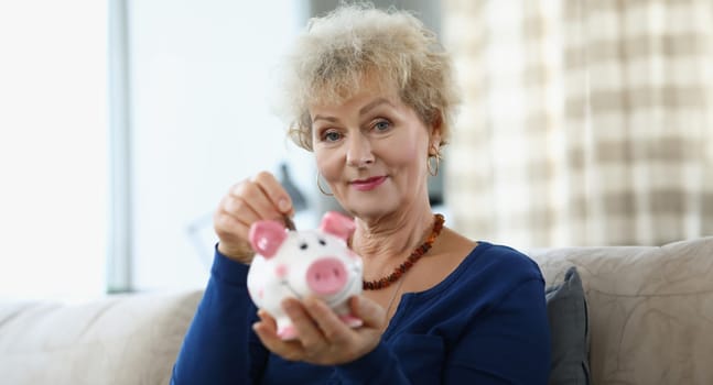 Elderly woman putting coin into piggy bank at home. Saving on retirement concept