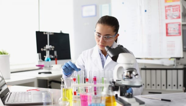 Chemist woman holding test tube with liquid in front of microscope in laboratory. Pharmaceutical industry concept