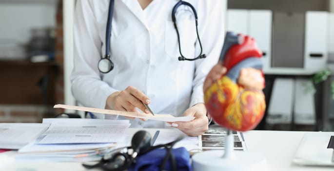 Doctor cardiologist examining cardiogram against background of artificial heart model closeup. Diagnosis of myocardial infarction and angina pectoris concept