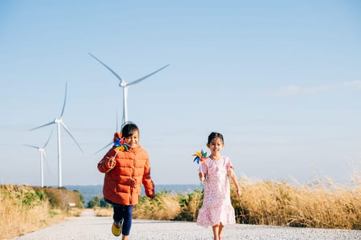 Two kids girl holding pinwheel joyfully run by windmills. Family near turbines embodies clean energy essence. Children happiness windmill circle against sun symbolizes sustainable cheerful community.