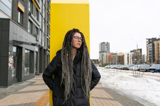 Street fashion concept, young European woman with dreadlocks dressed in a dark jacket walks through the city.