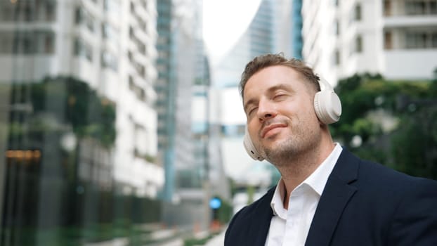 Caucasian businessman listen headphone and dancing to relaxing music with cheerful and motivation. Skilled project manager enjoy to listen relaxed song with blurred background at urban city. Urbane.