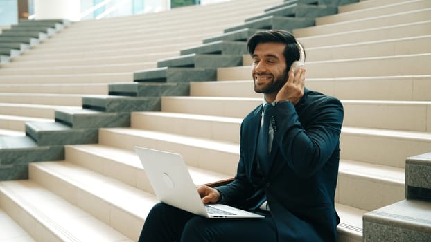 Professional business man sitting at stairs while working on laptop. Skilled project manager listening music from headphone and checking email and discussion about marketing plan. Outdoor. Exultant.